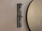Vertical Delsie Double Sconce, dark frame with two illuminated bulbs behind ribbed clear glass, mounted on a wall near a mirror.