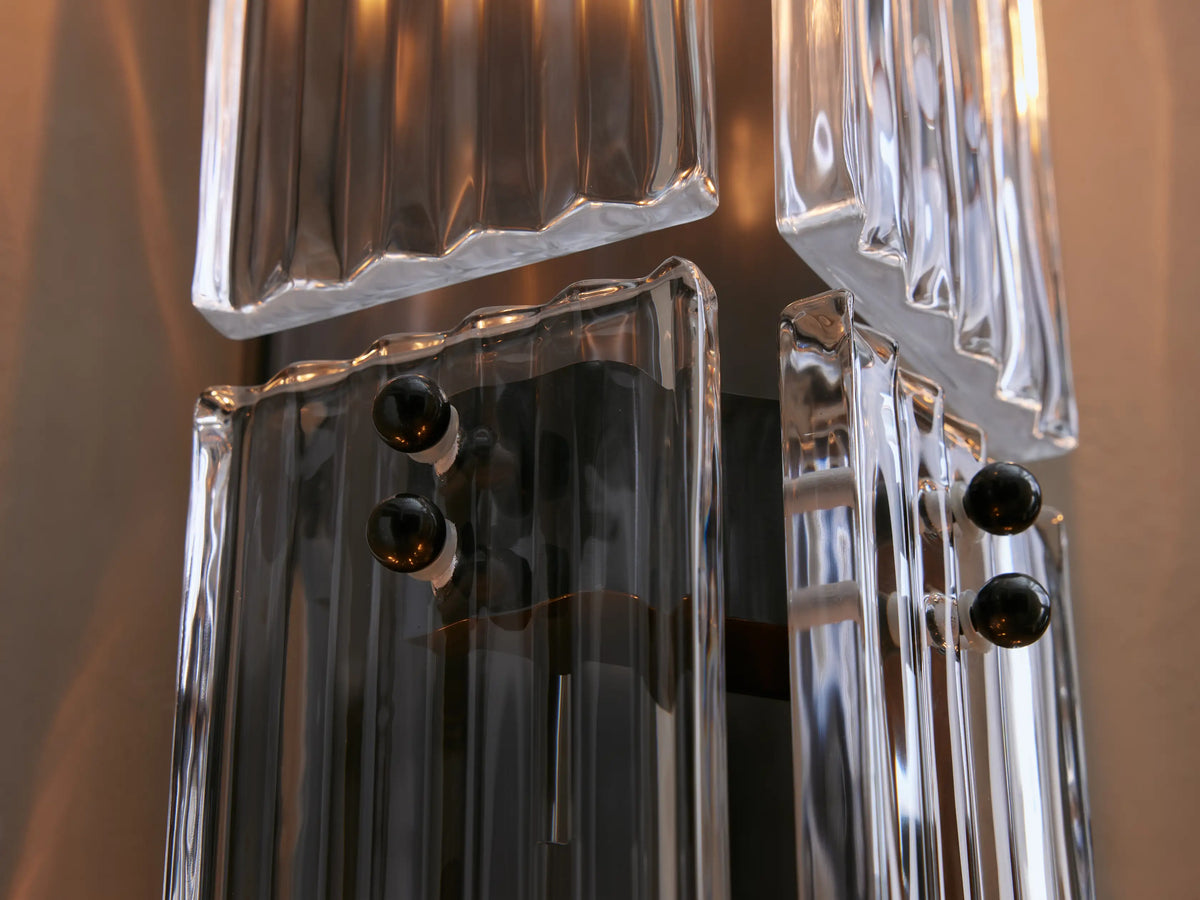 Vertical Delsie Double Sconce with fluted clear glass and dark bronze-toned frame, revealing bulbs, mounted on a light wall by a mirror.