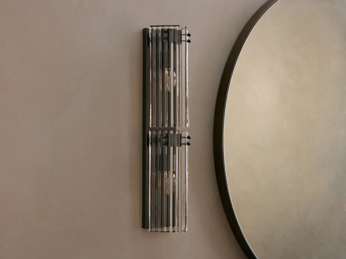 Delsie Double Sconce with two illuminated bulbs and fluted glass shades, mounted vertically on a light wall next to a round mirror.