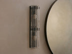 Delsie Double Sconce with two illuminated bulbs and fluted glass shades, mounted vertically on a light wall next to a round mirror.
