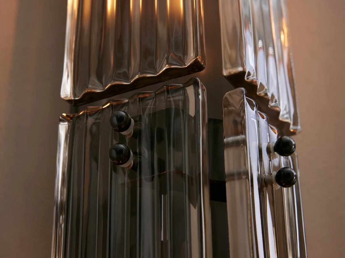 Delsie Double Sconce, vertical, clear fluted glass panels, dark bronze base, visible bulbs, on a light wall beside a round mirror.