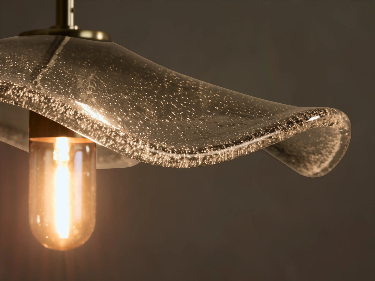 Close-up of a clear seeded glass wavy pendant light with a warm glowing bulb, suspended by a brass rod.