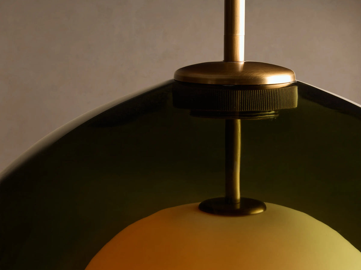 Front view of the Romy Pendant, featuring a dark green glass outer shade, an illuminated spherical cream inner diffuser, and a brass rod.