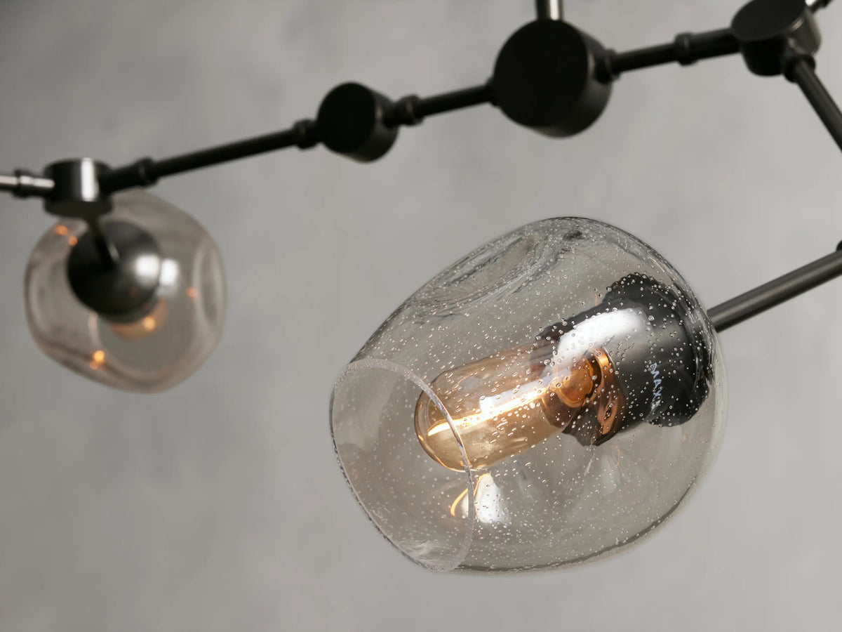 Noella Bronze Chandelier with bronze-finished steel frame and clear glass globes, featuring black arms and bulbs. Mounted on light gray wall.