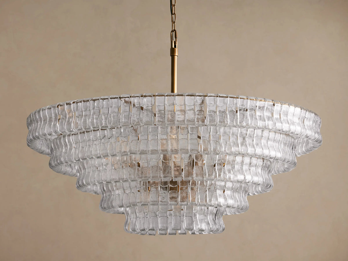 Ghiaccio Round Chandelier with tiered, curved and textured clear glass panels, brass chain, against a light brown wall.
