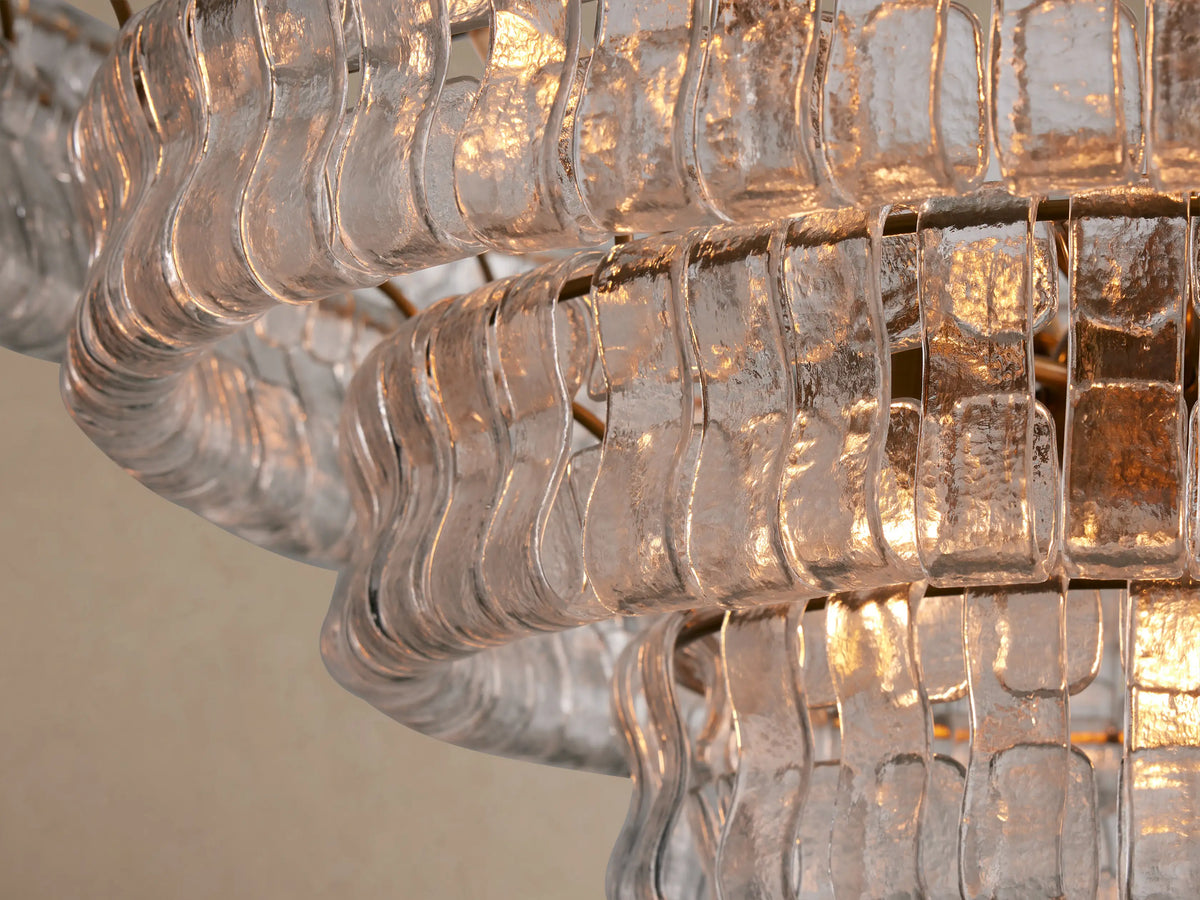 Ghiaccio Round Chandelier close-up, showing wavy, textured clear glass pieces arranged in tiers, illuminated with a warm light.