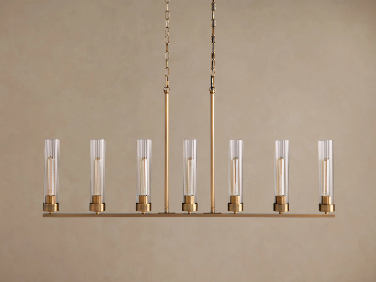 Andreas Linear Chandelier: Plated brass finish, 7 clear ribbed glass shades with visible filament bulbs, hanging from two chains.