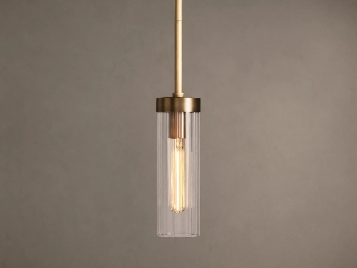 Brass Andreas Pendant with clear ribbed glass shade & illuminated filament bulb, hanging vertically against a neutral wall.