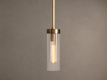 Brass Andreas Pendant with clear ribbed glass shade & illuminated filament bulb, hanging vertically against a neutral wall.