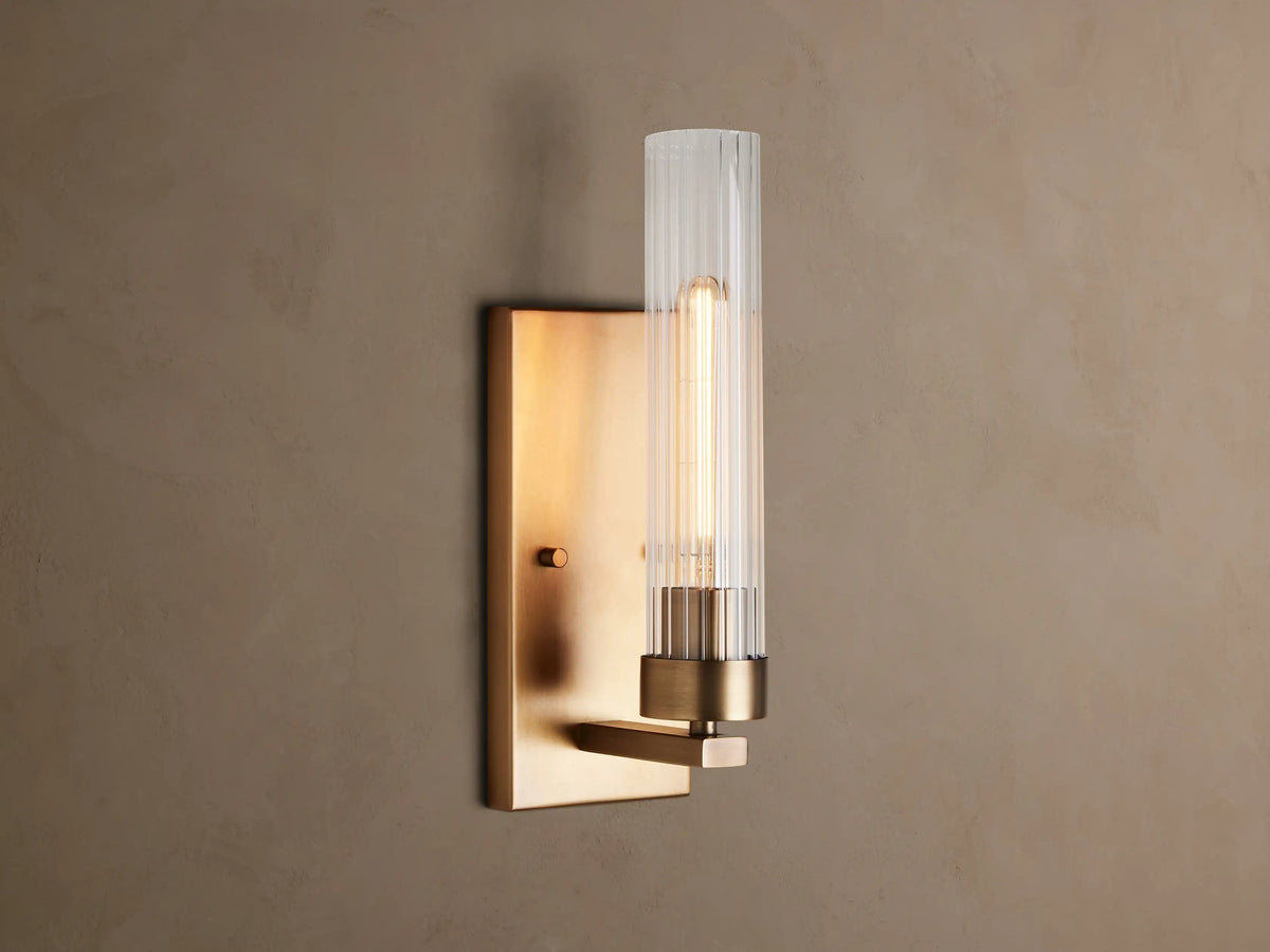 Andreas Sconce: Brass wall light with a rectangular backplate, clear fluted cylindrical glass shade, and a glowing filament bulb.