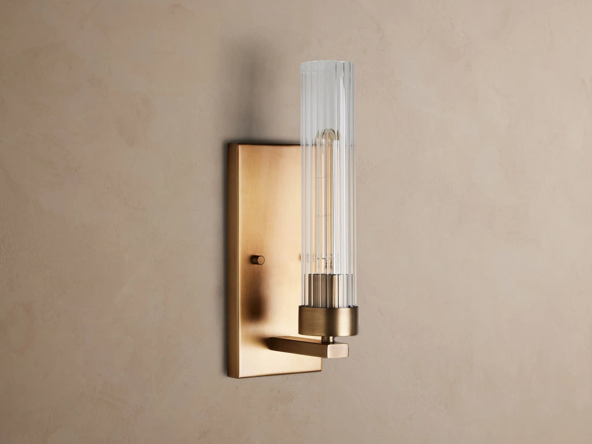 Andreas Sconce: Brass wall light with a rectangular backplate, clear fluted cylindrical glass shade, and a glowing filament bulb.