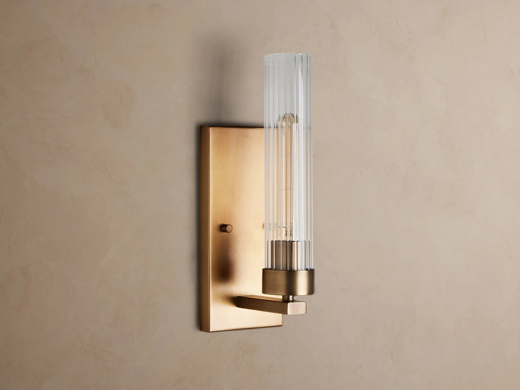 Andreas Sconce: Brass wall light with a rectangular backplate, clear fluted cylindrical glass shade, and a glowing filament bulb.