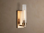 Andreas Sconce: Brass wall light with a rectangular backplate, clear fluted cylindrical glass shade, and a glowing filament bulb.