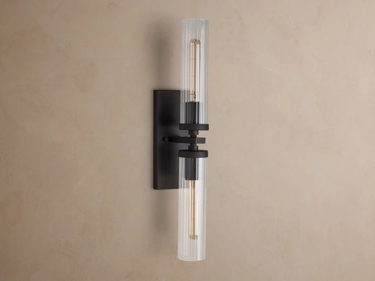 Andreas Double Sconce with two clear fluted glass shades, a dark bronze frame, and glowing exposed filament bulbs.