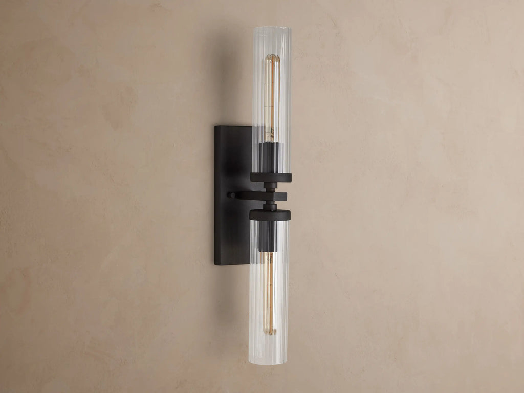 Andreas Double Sconce with two clear fluted glass shades, a dark bronze frame, and glowing exposed filament bulbs.