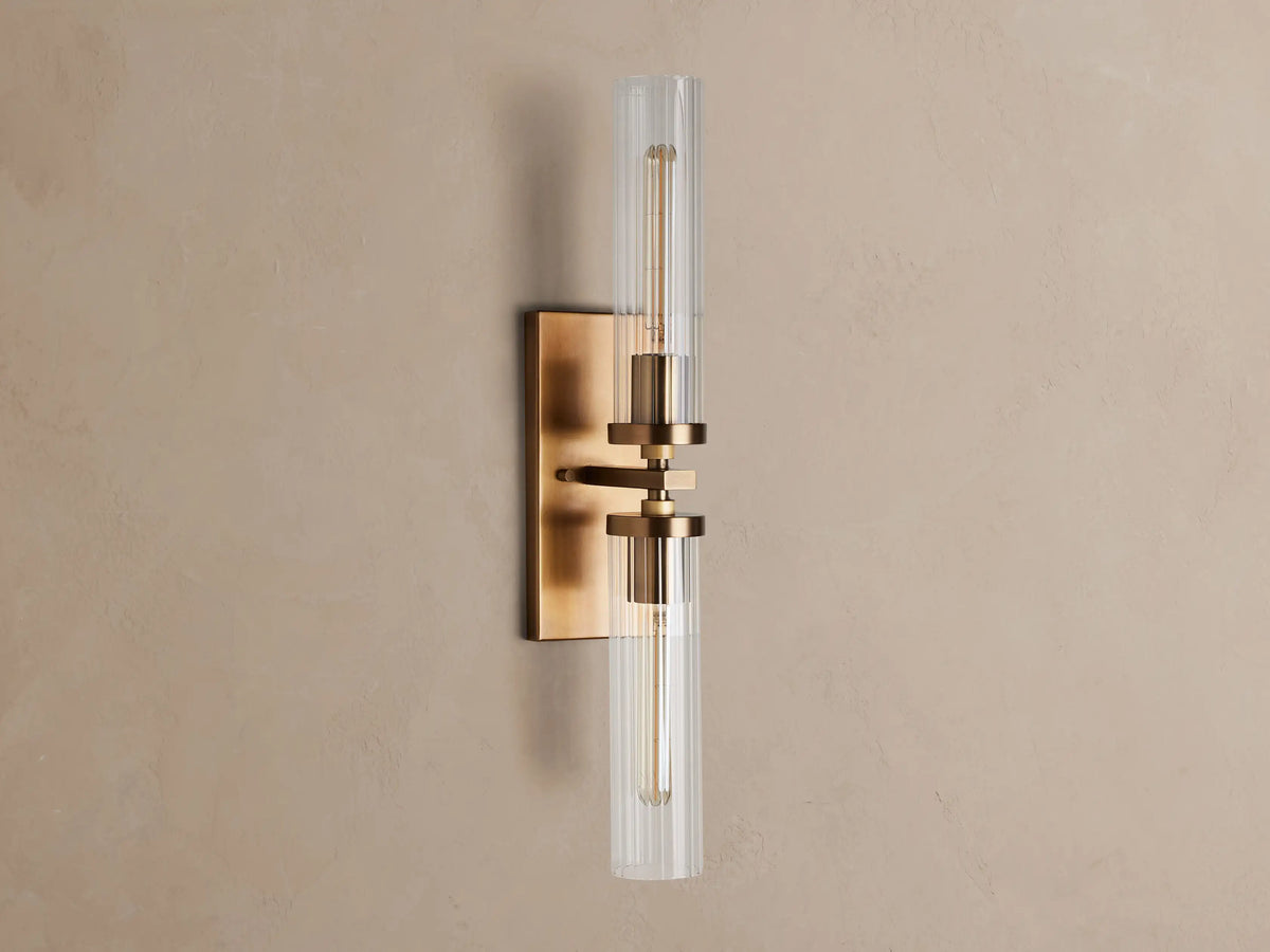 Andreas Double Sconce: rectangular brass backplate, two glowing T9 bulbs inside clear ribbed glass cylinders, mounted on a warm grey wall.