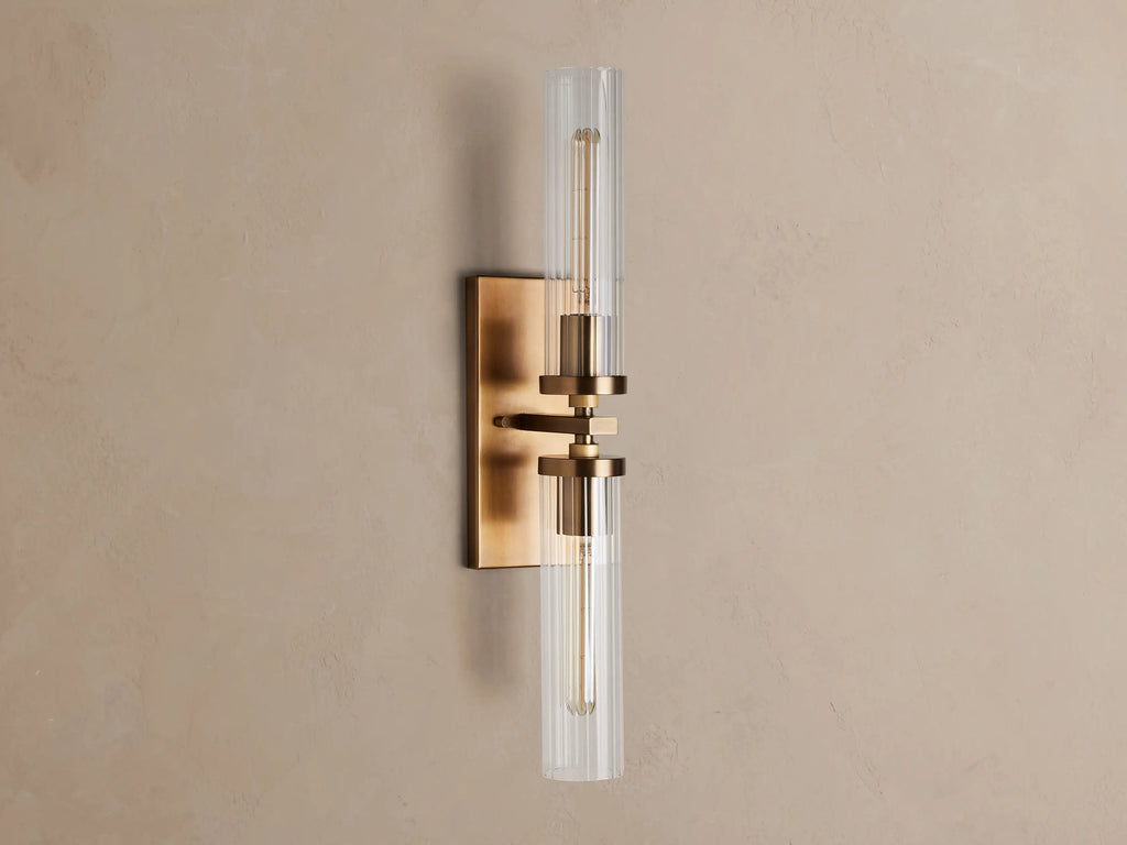 Andreas Double Sconce: rectangular brass backplate, two glowing T9 bulbs inside clear ribbed glass cylinders, mounted on a warm grey wall.