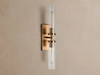 Andreas Double Sconce: rectangular brass backplate, two glowing T9 bulbs inside clear ribbed glass cylinders, mounted on a warm grey wall.