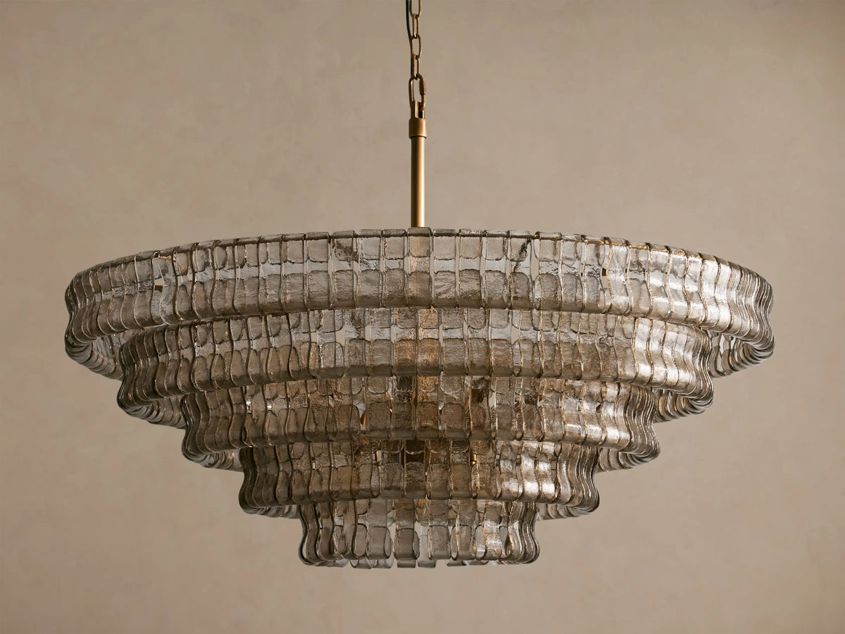 Ghiaccio Round Chandelier featuring tiered, textured smoky glass layers, suspended by a brass chain against a neutral wall.