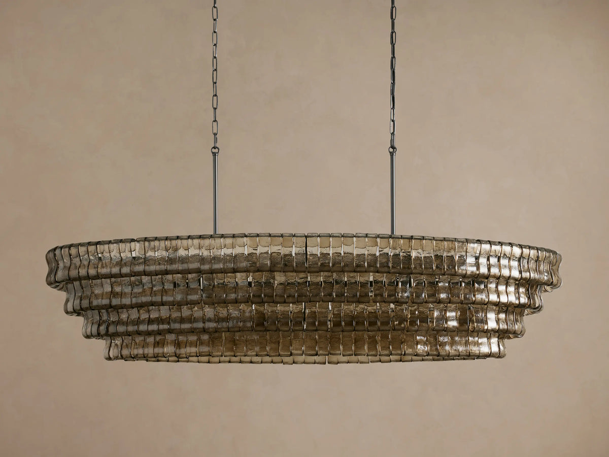 Ghiaccio Oval Chandelier, tiered with textured, curved smoke glass, glowing warmly, suspended by chains against a neutral wall.