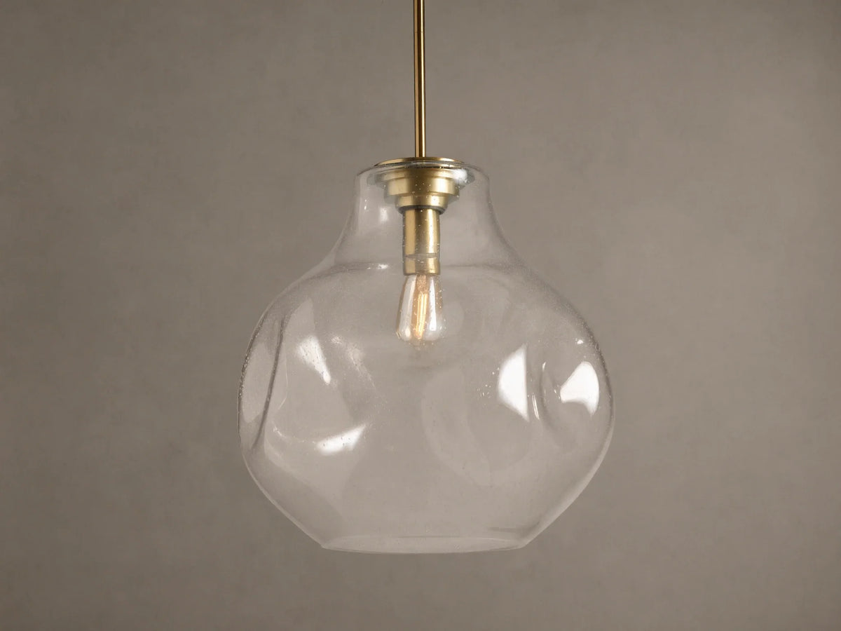 Organic 16" Pendant: Clear, seeded mouth-blown glass shade, brass stem, glowing filament bulb, against a brown wall.