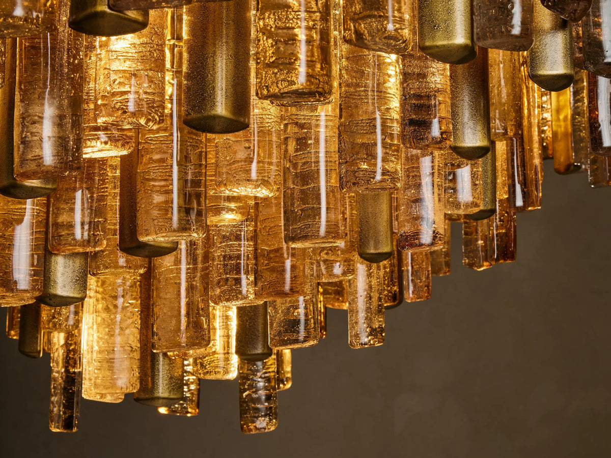 Close-up of Neve Chandelier: numerous amber and brass cylindrical elements, some clear glass, glowing warmly from within.