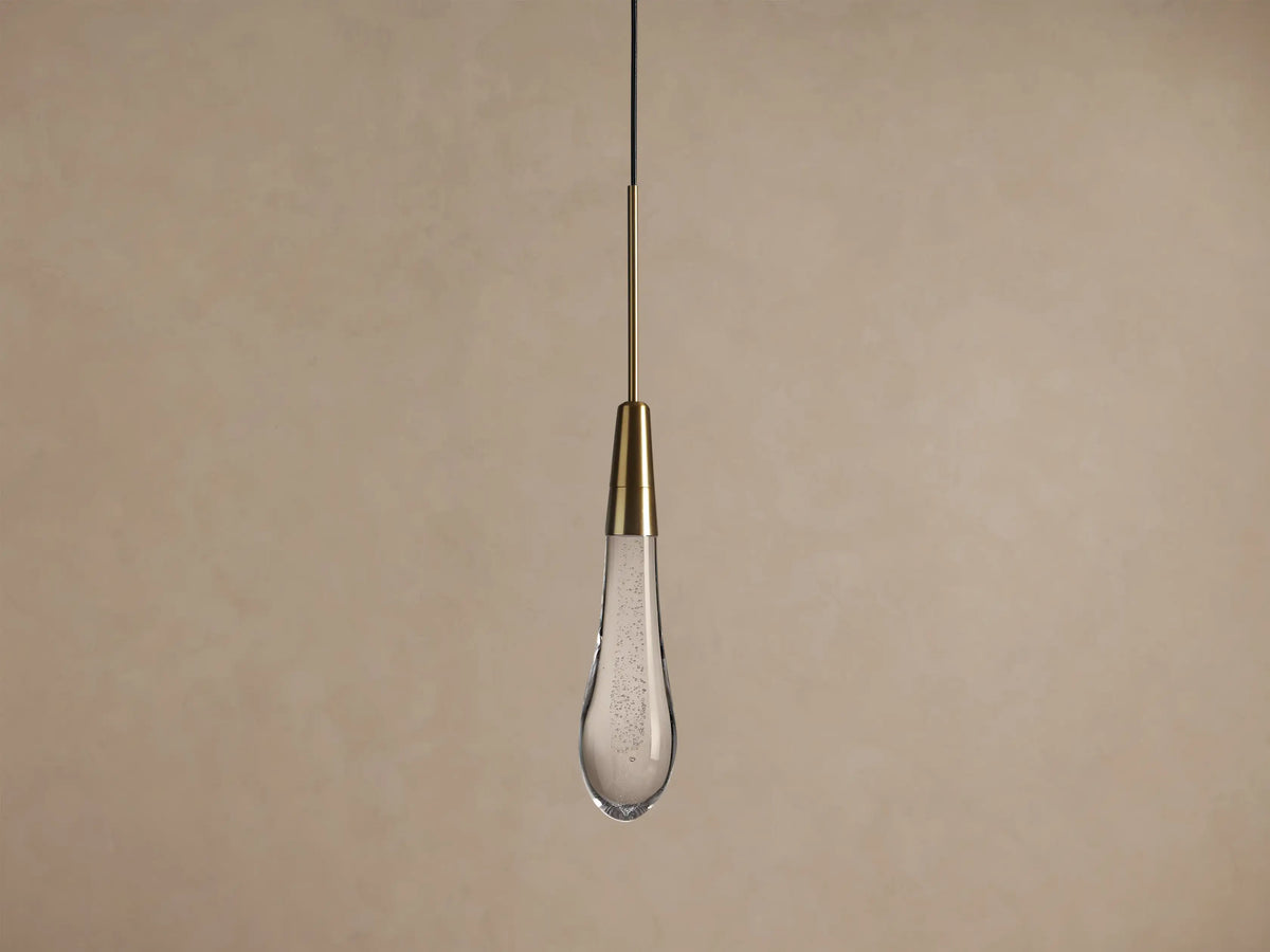 Soltaire Pendant: illuminated clear glass teardrop with internal bubbles & brass neck, hanging from a black cord against a neutral wall.