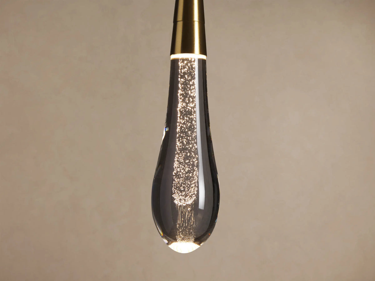 Soltaire Pendant: teardrop clear glass pendant with bubbles, brass top, hanging vertically against a plain wall.