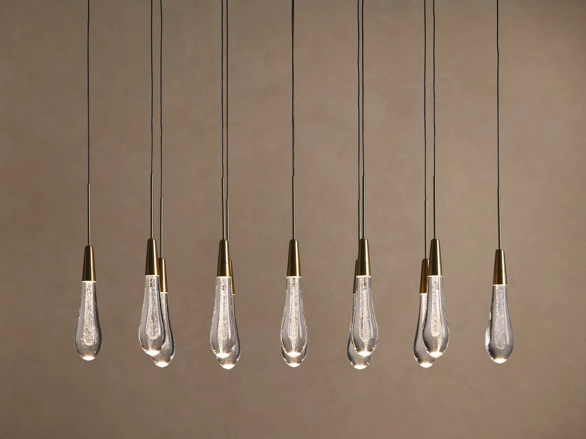 Soltaire Linear Chandelier with 8 clear, tear-drop glass pendants featuring brass tops & internal bubbles, glowing warmly.