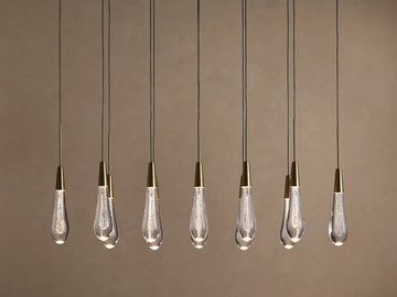 Soltaire Linear Chandelier with 8 clear, tear-drop glass pendants featuring brass tops & internal bubbles, glowing warmly.