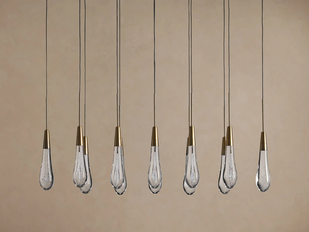Soltaire Linear Chandelier: Multiple clear bubbled teardrop glass pendants with conical brass tops, suspended by thin black cords.