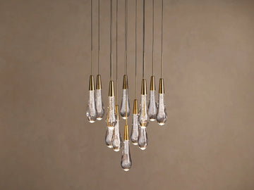 Soltaire chandelier with many illuminated teardrop glass pendants, brass caps, and internal bubbles, clustered against a neutral wall.