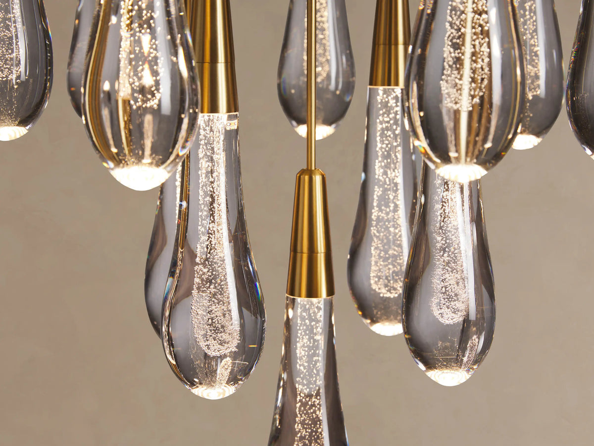 Close-up of Soltaire Round Chandelier showing multiple clear, bubbly teardrop glass pendants with brass tops, glowing brightly.
