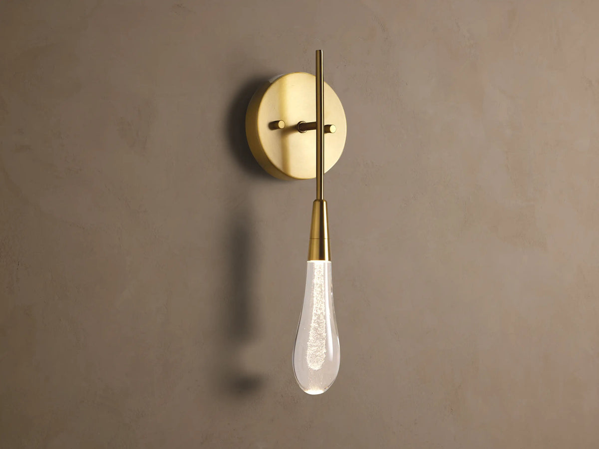 Soltaire Sconce: Round brass backplate, vertical arm, and glowing teardrop glass shade with internal bubbles mounted on a wall.