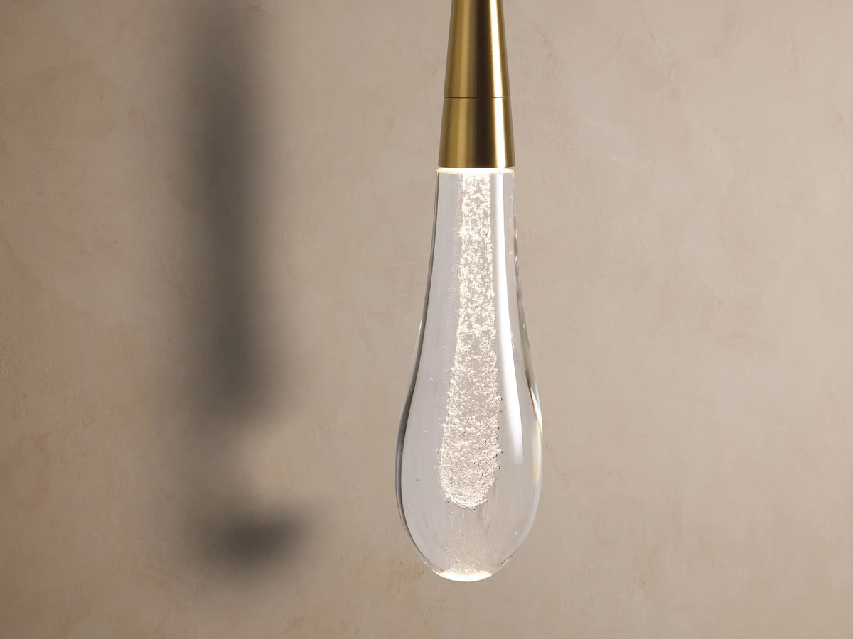 Soltaire Sconce: Round brass wall plate with a vertical rod and a clear, bubble-filled glass teardrop shade, mounted on a textured wall.