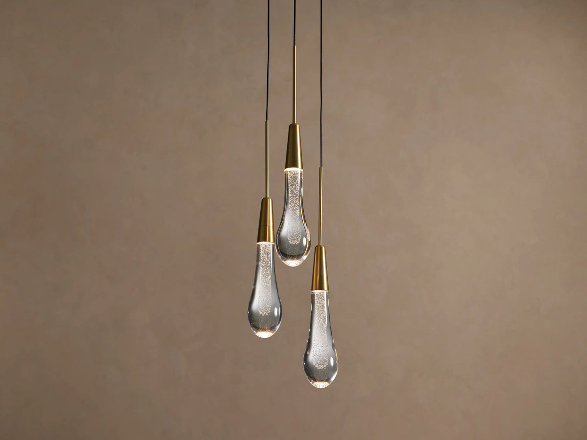 Three Soltaire Cluster Pendants with brass conical tops and clear, bubbly glass teardrop shades hang illuminated against a tan wall.