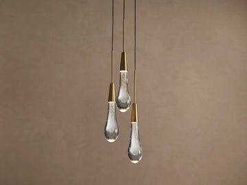 Three Soltaire Cluster Pendants with brass conical tops and clear, bubbly glass teardrop shades hang illuminated against a tan wall.