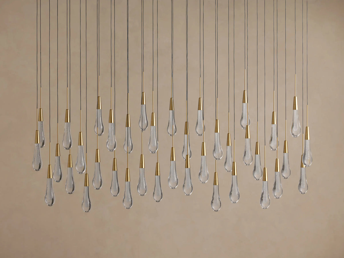 Soltaire Linear Chandelier with many clear teardrop glass pendants, brass caps, hanging at varying heights on a beige background.