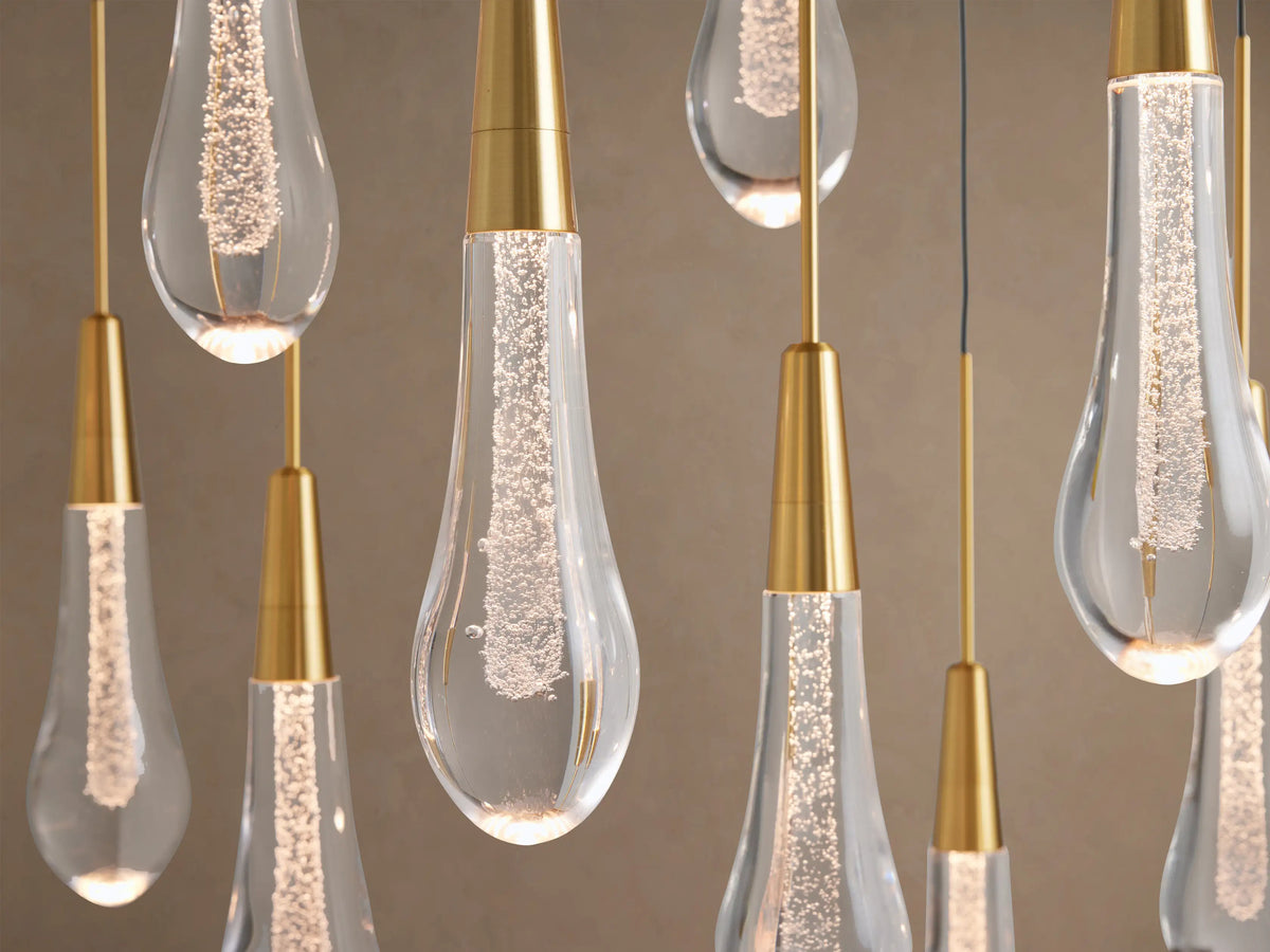 Soltaire Linear Chandelier, close-up of glowing clear glass teardrop pendants with brass caps & embedded shimmering lights.