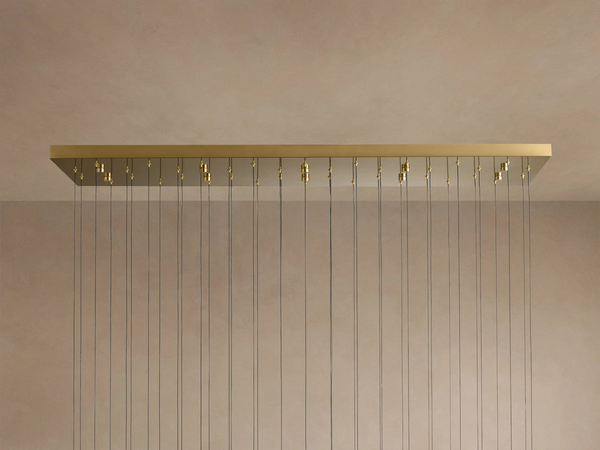 Soltaire Linear Chandelier: long brass rectangular canopy with many thin cords hanging down against a light beige wall.