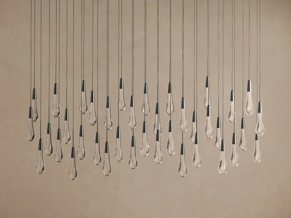 Soltaire Linear Chandelier with numerous clear tear-drop glass pendants, dark caps, suspended at varying heights against a light wall.