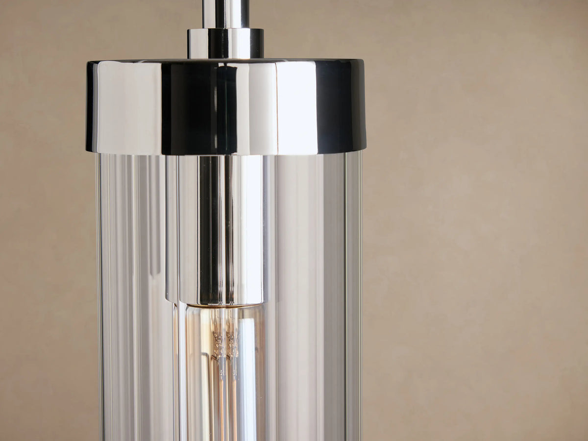 Polished nickel Andreas Pendant, featuring a clear glass cylinder shade enclosing an Edison filament bulb, suspended against a light brown wall.