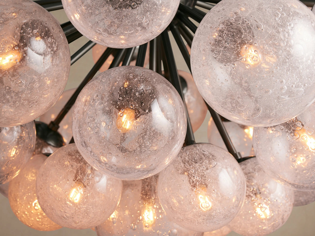 Close-up of Mandoza Chandelier's many clear, seeded glass globes with warm glowing LED filament bulbs on a dark metal frame.