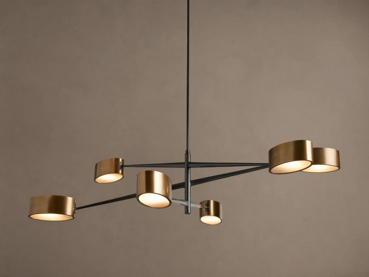 Etoile Chandelier: Modern design with black arms and six brass cylindrical shades glowing warmly, suspended against a brown wall.