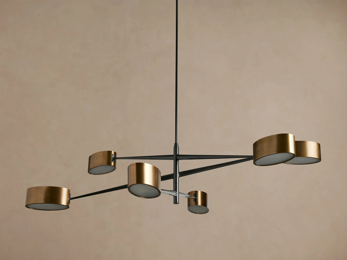 Etoile Chandelier: Modern design with black arms and six brass cylindrical shades glowing warmly, suspended against a brown wall.