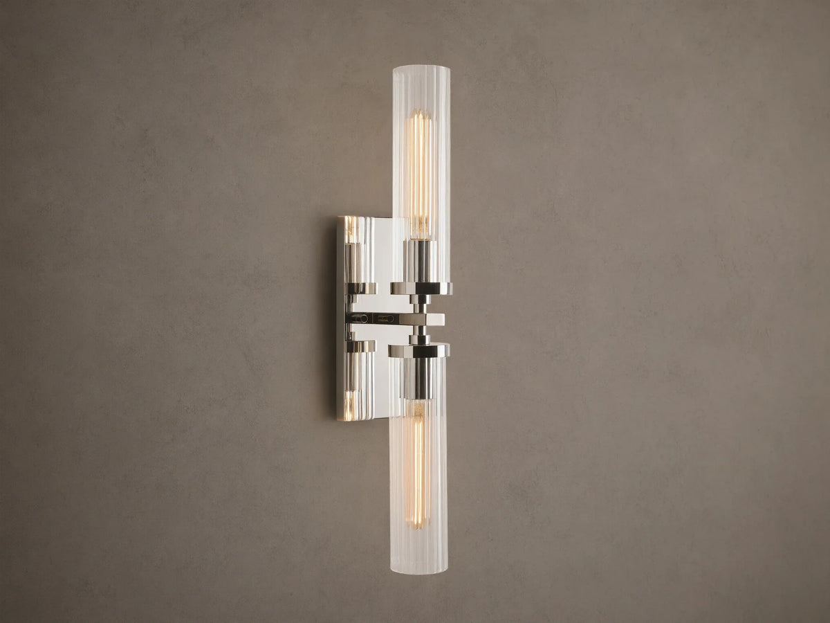 Andreas Double Sconce: vertical polished nickel hardware, two ribbed clear glass shades, warm glowing bulbs, mounted on a taupe wall.