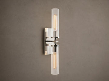 Andreas Double Sconce: vertical polished nickel hardware, two ribbed clear glass shades, warm glowing bulbs, mounted on a taupe wall.
