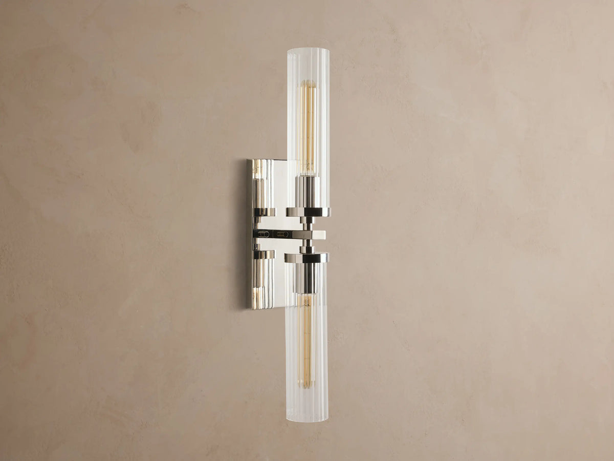 Andreas Double Sconce: vertical polished nickel hardware, two ribbed clear glass shades, warm glowing bulbs, mounted on a taupe wall.