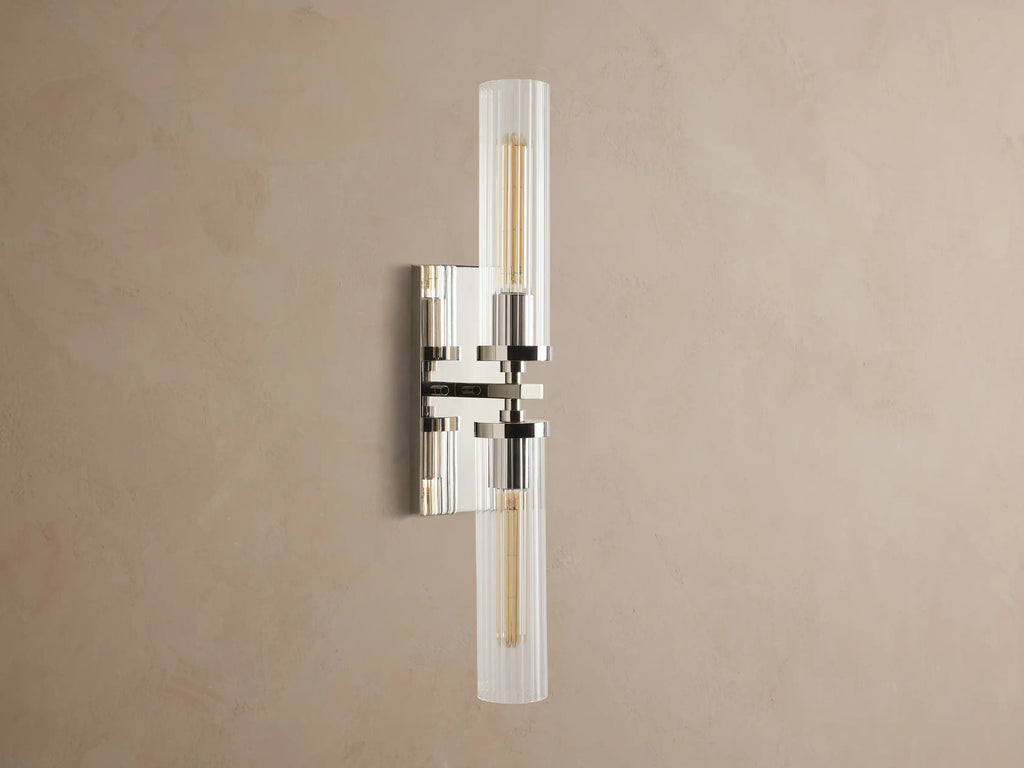 Andreas Double Sconce: vertical polished nickel hardware, two ribbed clear glass shades, warm glowing bulbs, mounted on a taupe wall.