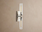 Andreas Double Sconce: vertical polished nickel hardware, two ribbed clear glass shades, warm glowing bulbs, mounted on a taupe wall.